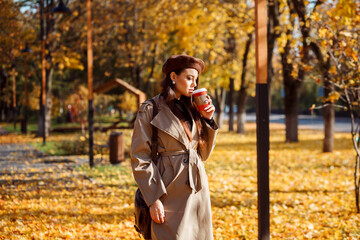 Attractive stylish woman is drinking coffee in the park in autumn, yellow leaves backgrouund.
