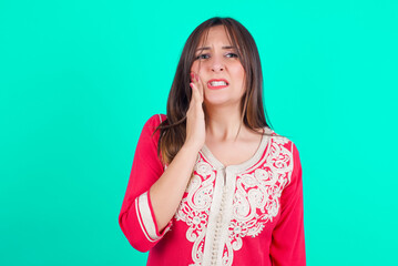 young beautiful moroccan woman wearing traditional caftan dress over green background touching mouth with hand with painful expression because of toothache or dental illness on teeth.