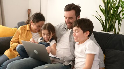 Cheerful happy family of four using laptop sitting on the sofa in cozy apartment, parents and two kids watching movie on computer, comedy, spends time on the weekend together at home - Powered by Adobe