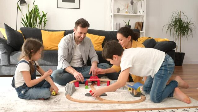 Lovely family of four spend weekend together at home, parents and two kids playing toys, sibling building railway, have a fun. Mom, dad and children sitting on floor in cozy living room and playing