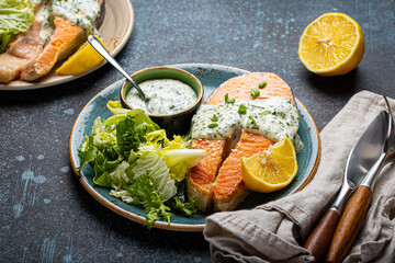 Healthy food meal cooked grilled salmon steak with white dill sauce and green salad leafs on plate on rustic concrete stone background table angle view, diet healthy nutrition dinner copy space