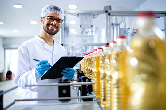 Portrait of an experienced caucasian technologist or worker standing by the production line conveyor machine with bottles of sunflower oil in food factory.