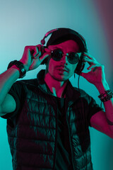 Hipster man with sunglasses and cap listen to music in headphones in studio with colorful pink and blue light. Creative colored male portrait