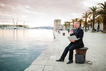 lachender Geschäftsmann mit Laptop sitzt auf Altstadtpromenade