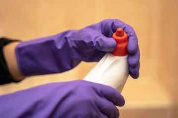 Hands in purple rubber gloves hold a plastic bottle of detergent and unscrew the red cap