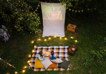 High angle view of children sitting on plaid and looking movie on backyard. Summer outdoor weekend activities with kids. Open air cinema.