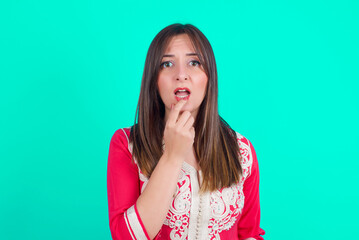 Nervous puzzled young beautiful moroccan woman wearing traditional caftan dress over green background opens mouth from surprise, reacts on sudden news.
