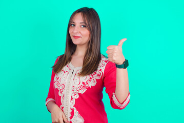 young beautiful moroccan woman wearing traditional caftan dress over green background doing happy thumbs up gesture with hand. Approving expression looking at the camera showing success.