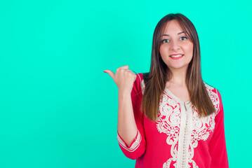 Lovely young beautiful moroccan woman wearing traditional caftan dress over green background pointing aside with forefinger, showing at copy space having news about bargains