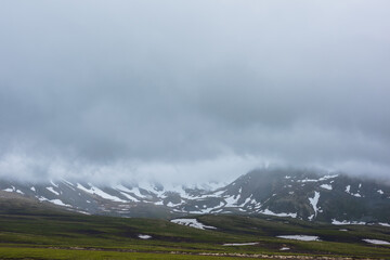 Dramatic alpine landscape with snowy mountains in gray low clouds. Bleak atmospheric scenery of tundra under lead gray sky. Gloomy minimalist view to mountain range among low rainy clouds in overcast.