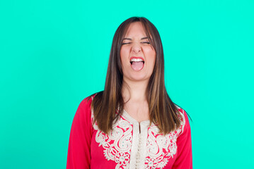 Fototapeta premium young beautiful moroccan woman wearing traditional caftan dress over green background sticking tongue out happy with funny expression. Emotion concept.