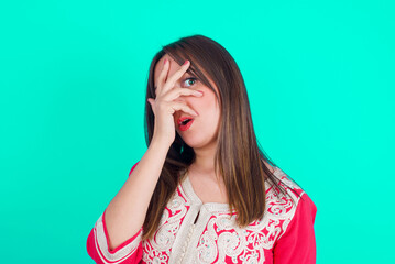 young beautiful moroccan woman wearing traditional caftan dress over green background peeking in shock covering face and eyes with hand, looking through fingers with embarrassed expression.