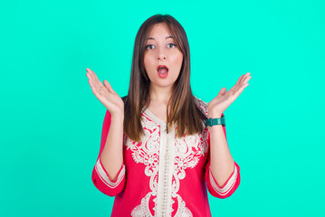 Surprised terrified young beautiful moroccan woman wearing traditional caftan dress over green backgrou Gestures with uncertainty, stares at camera, puzzled as doesn't know answer on tricky question, 