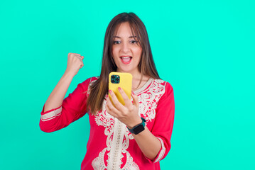 young beautiful moroccan woman wearing traditional caftan dress over green background holding in hands cell and rising his fist up being excited after reading good news.