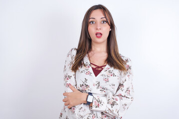 Shocked embarrassed young Arab woman wearing floral dress over white background  keeps mouth widely opened. Hears unbelievable novelty stares in stupor