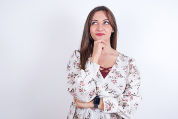 Thoughtful young Arab woman wearing floral dress over white background  holds chin and looks away pensively makes up great plan