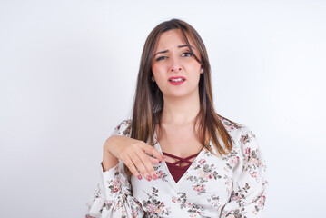 Portrait of dissatisfied young Arab woman wearing floral dress over white background  smirks face, purses lips and looks with annoyance at camera, discontent hearing something unpleasant