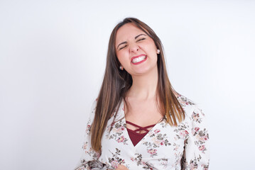 Positive young Arab woman wearing floral dress over white background   with overjoyed expression closes eyes and laughs shows white perfect teeth