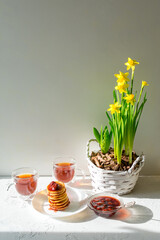 Fototapeta na wymiar Fritters with strawberry jam, two cups of tea and a bouquet of yellow daffodils in a basket on a white background.