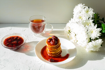 Fritters in a white plate with strawberry jam, a cup of tea and a bouquet of white chrysanthemums on a white table.