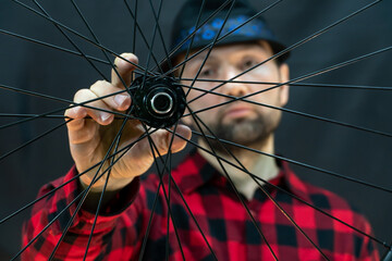 A fashionable bicycle mechanic in a red shirt and a black hat holds a wheel in his hand. Assembling wheels from a carbon rim using professional equipment for tension spokes. Bicycle repair.