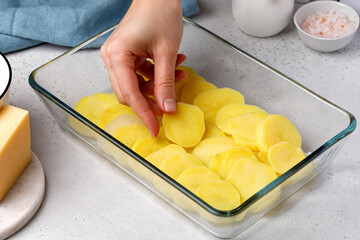 Step-by-step recipe of potato gratin. Hands stack potato slices to a glass baking dish. Potato casserole