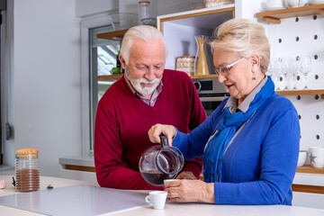 Happy senior couple in the kitchen. The wife prepares her husband his morning coffee, they are smiling and talking to each other.