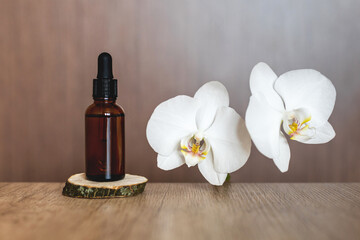 A glass bottle of serum with a pipette stands on a natural wooden podium with a white orchid on a brown textured table. Concept for advertising a product