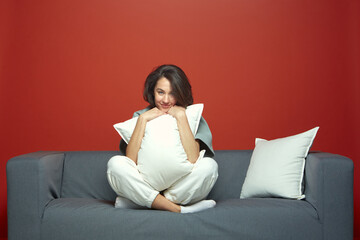 Obraz premium Pleased stylish young woman brunette hug white pillow sitting on cozy sofa, resting, relaxing on weekend