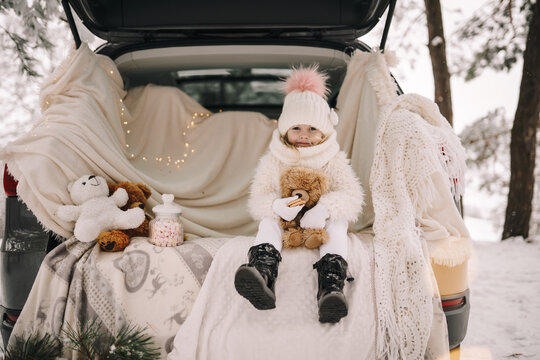 Little girl in the car. Little girl in white warm clothes. Soft teddy bear. Vacation in the winter forest.