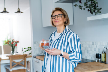Portrait of middle age smiling woman holding her mobile phone and drinking tea while standing in the modern kitchen. Morning habits and rituals. Day planning, working from home.