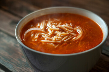 Hot Mexican noodle tomato soup with onion garlic and other condiment, in a white bowl, over an old wooden table, side view macro photography.