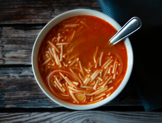 Hot Mexican noodle tomato soup with onion garlic and other condiment, in a white bowl, over an old wooden table, lay flat top view