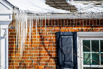 Large icicles hanging from snow covered roof of brick house indicating poor roof insulation. - obrazy, fototapety, plakaty