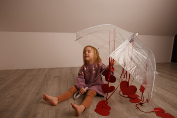 a beautiful blonde girl in a pink blouse with sequins is sitting on the floor with her eyes closed, and next to her is an umbrella with red hearts