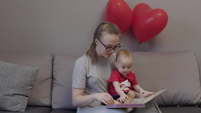 young caucasian mother in glasses reading education book to her baby child kid in red t-shirt. mother and toddler sitting grey sofa in living room looking picture book with red heart balloons behind