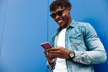 happy curly african american man wearing sunglasses using mobile phone and smiling against blue...