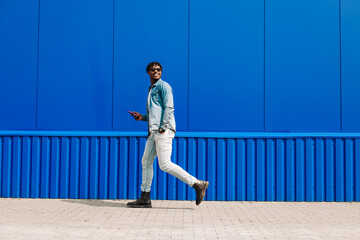 stylish African American man in full growth, uses a smartphone and walks past a blue wall while going to work