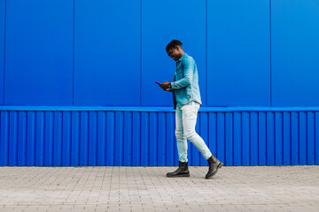stylish African American man in full growth, uses a smartphone and walks past a blue wall while...