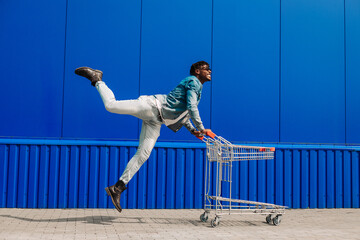 Excited happy african american man having fun while shopping, man pushing shopping cart while...