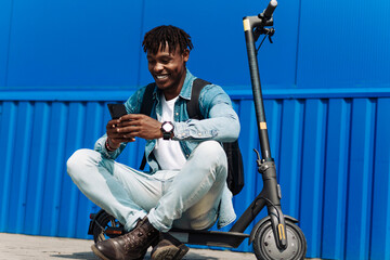 Stylish smiling african american man sitting on his electric scooter near a large building, using a...