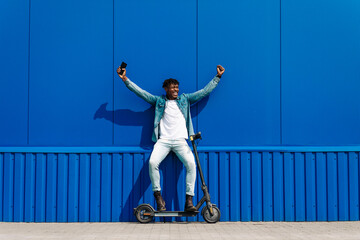 Excited African American man using an electric scooter on a city street. Man holding mobile phone,...