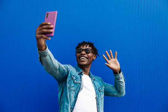 african american man in a casual shirt, on a blue background Takes a selfie on a mobile phone, making a greeting gesture