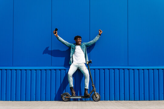 Excited African American man using an electric scooter on a city street. Man holding mobile phone, smiling, feeling joy