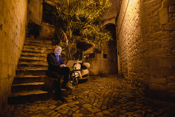 glücklich lachender Geschäftsmann mit Laptop sitzt in Altstadt-Gasse auf Stein-Treppe, Nacht