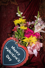 Valentine flowers and heart that says Happy Valentine's Day against dark draped background.