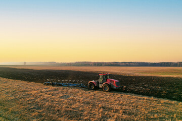 Tractor Plowing field on sunset. Red Tractor with Plough on Plowed. Ploughing and Soil Tillage. Agricultural Tractor on Cultivation Field for Sowing Seeds. Big Tractor During Field Cultivating. .
