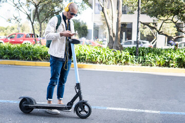 Latin man with glasses taking a scooter with his cell phone and headphones on a city street © FABIAN PONCE GARCIA