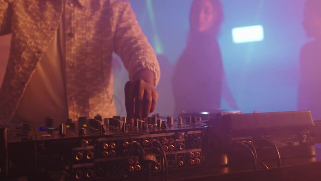 Close-up of technic equipment, DJ controlling mixing desk. Tilt up reveal dancing people. Nightlife, modern music and entertainment concept.