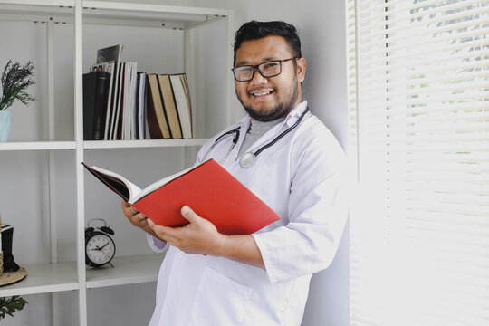 Smiling Male Doctor Looking At Camera Leaning Back And Reading Medical Book In Hospital